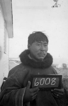 A black-and-white photograph of an Inuit guy holding a tiny chalkboard because of the number 6008.